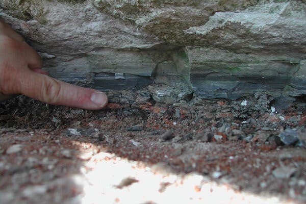 Una vista de cerca de un dedo humano apuntando a una capa de sedimento gris oscuro en la roca sobre una capa rojiza.